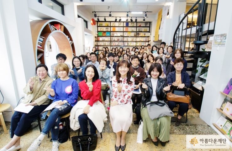 행사에 참석한 참석자들이 의자에 앉아서 손으로 하트를 그리며 정면을 바라보고 있다
