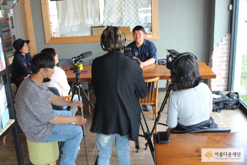 카페 <이매진>의 찰스와 인터뷰하고 있는 활동가들, 중앙에 테이블 뒤에 찰스가 있고, 그 뒤로 촬영을 하는 카메라와 활동가가 중앙과 오른쪽에, 마이크와 음향을 담당하는 활동가는 왼쪽에 있다.
