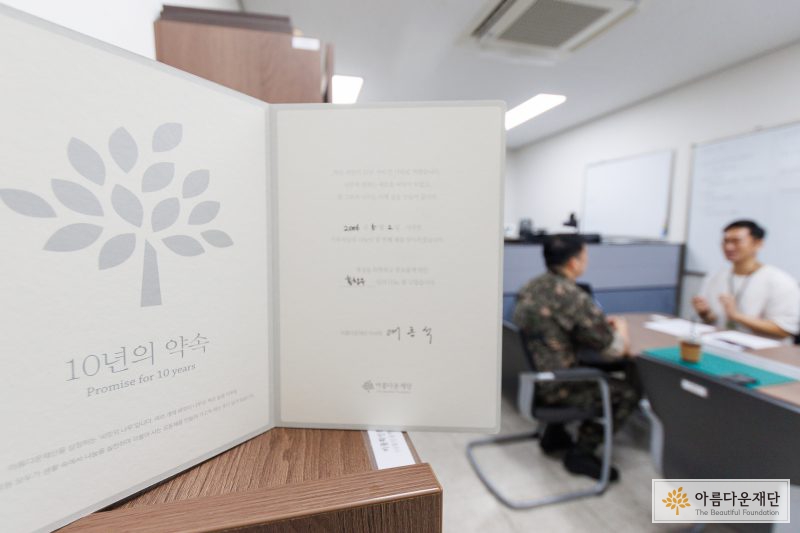 기부 10년차 감사패 이미지. 좌측으로 아름다운재단 씨앗나무 로고가 있고 오른쪽은 감사메세지가 적혀 있다. 감사패 뒷편으로는 인터뷰 중인 홍창우 기부자가 보인다.