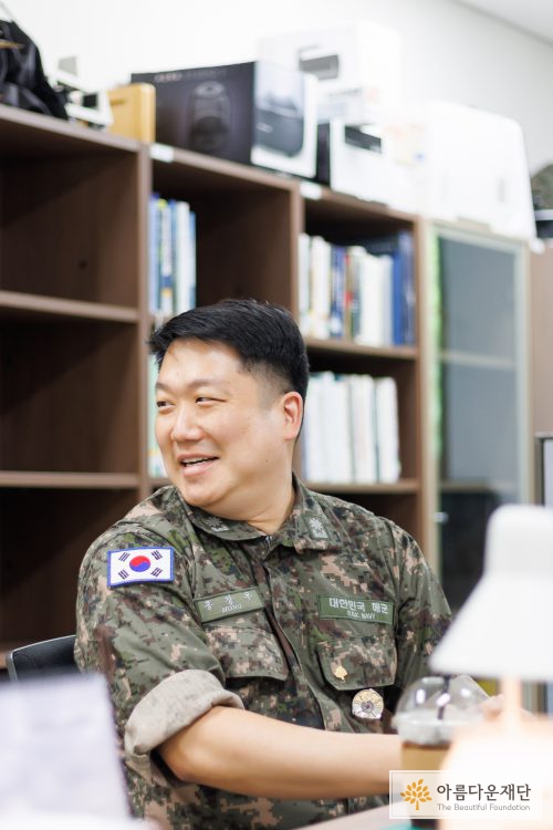 기부 10년차 감사장이 진열되어 있는 책장 쪽으로 뒤 돌아 보면서 웃고 있는 홍창우 기부자