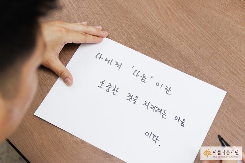 기부자가 백지 위에 나눔 한 마디를 쓰고 있다. 기부자는 "나에게 나눔이란 소중한 것을 지키려는 마음" 이라고 썼다.