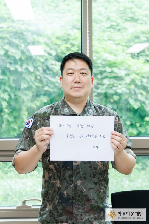 홍창우 기부자가 자신이 쓴 나눔 한 마디 메세지 보드를 들고 밝게 웃으며 촬영하고 있다.