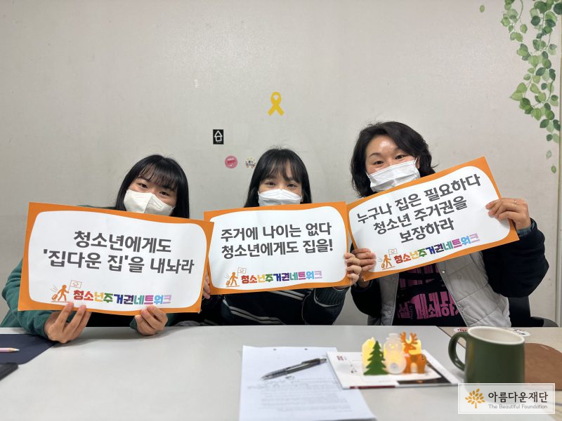 사진의 왼쪽부터 청소년주거권네트워크의 김시연, 정찬송, 변미혜 활동가가 책상 앞 의자에 앉아 피켓(청소년에게도 집다운집을 내놔라, 주거에 나이는 없다 청소년에게도 집을, 누구나 집은 필요하다 청소년 주거권을 보장하라)을 들고 있다. 세 사람 모두 마스크를 쓰고 있다.