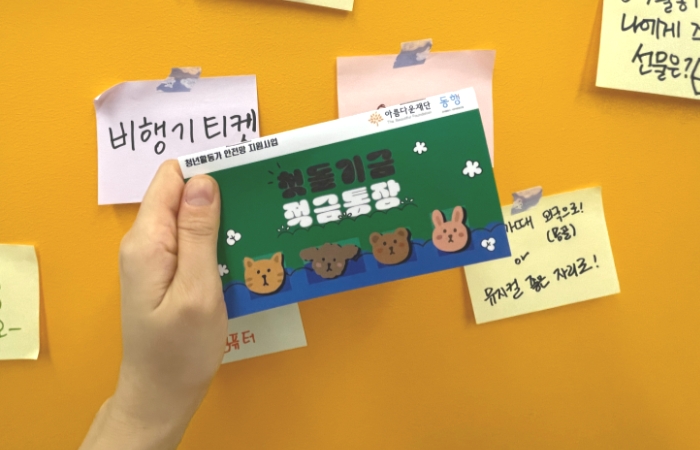 공익활동 1주년 나에게 주고싶은 선물을 적은 포스트잇이 붙어있고, 앞에는 첫돌기금 적금통장을 들고 있는 손이 보인다.