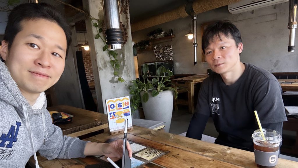 남성 두명이 카페에 앉아 사진을 찍고 있다. 