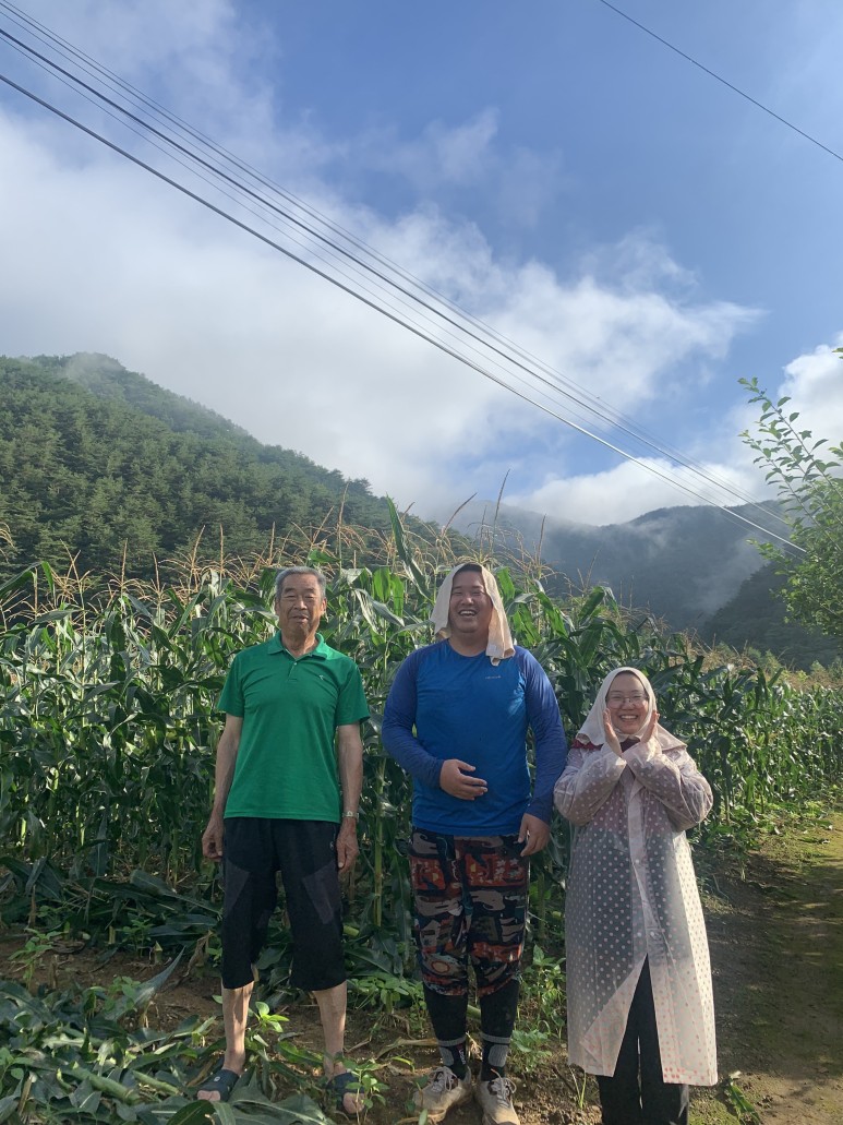 세 명이 옥수수 밭 앞에 서 있다. 맨 왼쪽에 어르신 농부, 중간에는 청년 남성, 오른쪽에는 청년 여성이 있다.