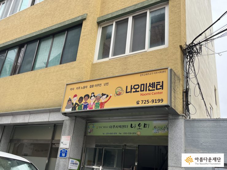 노란 건물 입구에 나오미센터 간판이 달려있고, 간판에는 다양한 인종과 복장을 한 사람들이 서 있다.