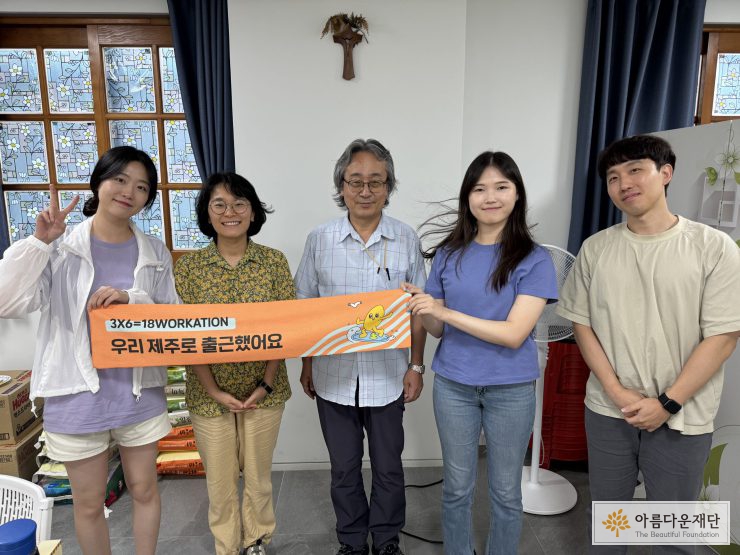 김상훈 사무국장이 가운데 서 있고, 양옆으로 아름다운재단 매니저가 서 있다. 현수막을 들고 있고, 현수막에는 우리 제주로 출근했어요! 라고 써 있다.