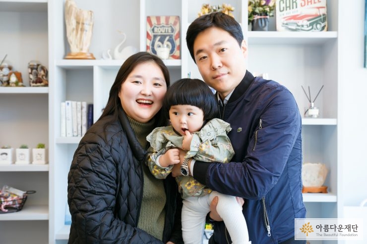 긴 생머리에 검정색 옷을 입은 엄마와 남색 자켓을 입은 아빠 그리고 연두색 원피스를 입은 딸 세 가족이 정면을 보며 찍은 사진