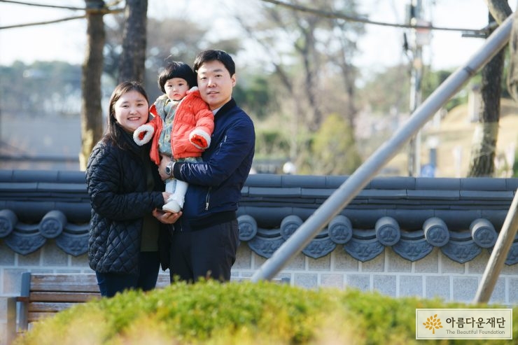 검은색 옷을 입은 엄마와 남색 자켓을 입은 아빠 그리고 주황색 패딩을 입은 딸 아이가 아빠에게 안겨 셋이 찍은 가족사진