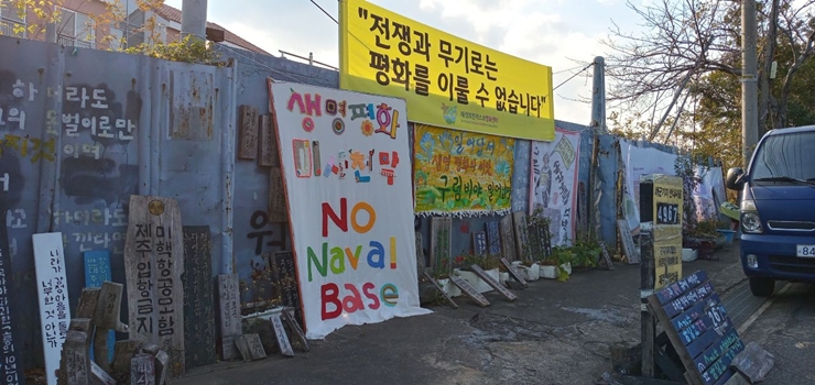 해군기지 공사장 정문에 '전쟁과 무기로는 평화를 이룰 수 없습니다' '생명평화 미사천막 NO Naval Base' 라고 적힌 현수막 두개와, 아래쪽으로 관련하여 여러 목소리를 내는 다양한 피켓이 놓여있다.