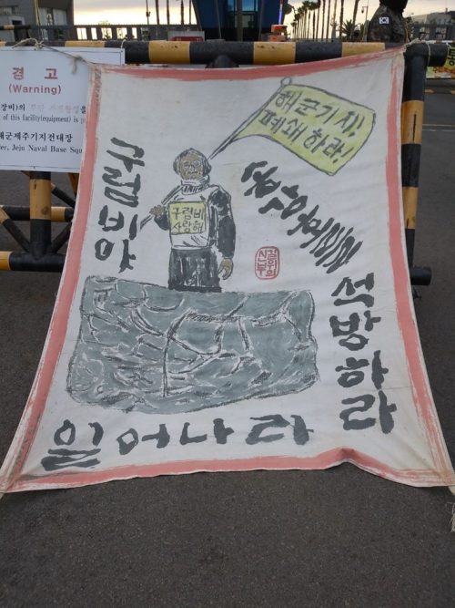 평화활동가 송강호 박사 석방 자보. 구럼비 위에 서서 '해군기지 폐쇄하라' 라는 깃발을 든 송강호 박사의 이미지와, 구럼비야 일어나라, 송강호를 석방하라는 문구가 적혀있다.