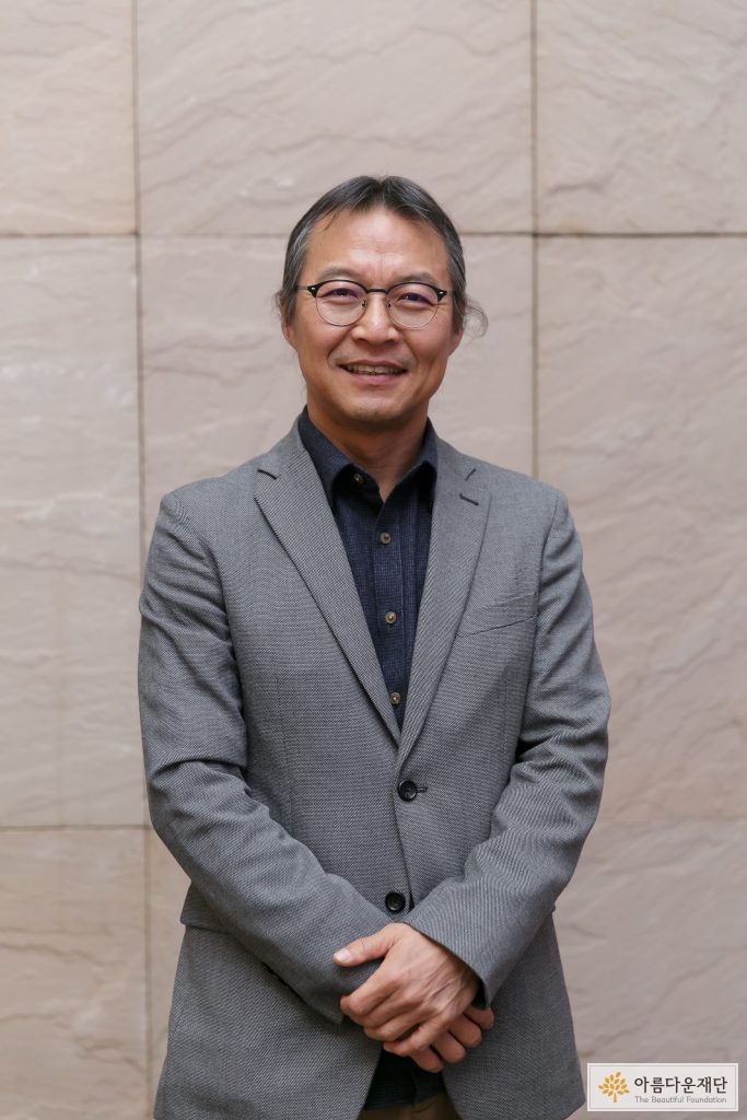 회색 자켓을 입고 안경을 쓴 머리를 묶은 남성이 두 손을 가지런히 모은 채 정면을 바라보며 서있음.