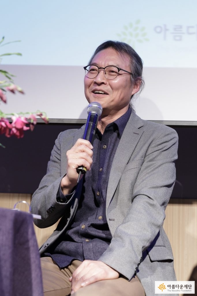 회색 자켓을 입고 안경을 쓴 남성이 앉아서 마이크를 들고 앞을 보며 말을 하고 있음.