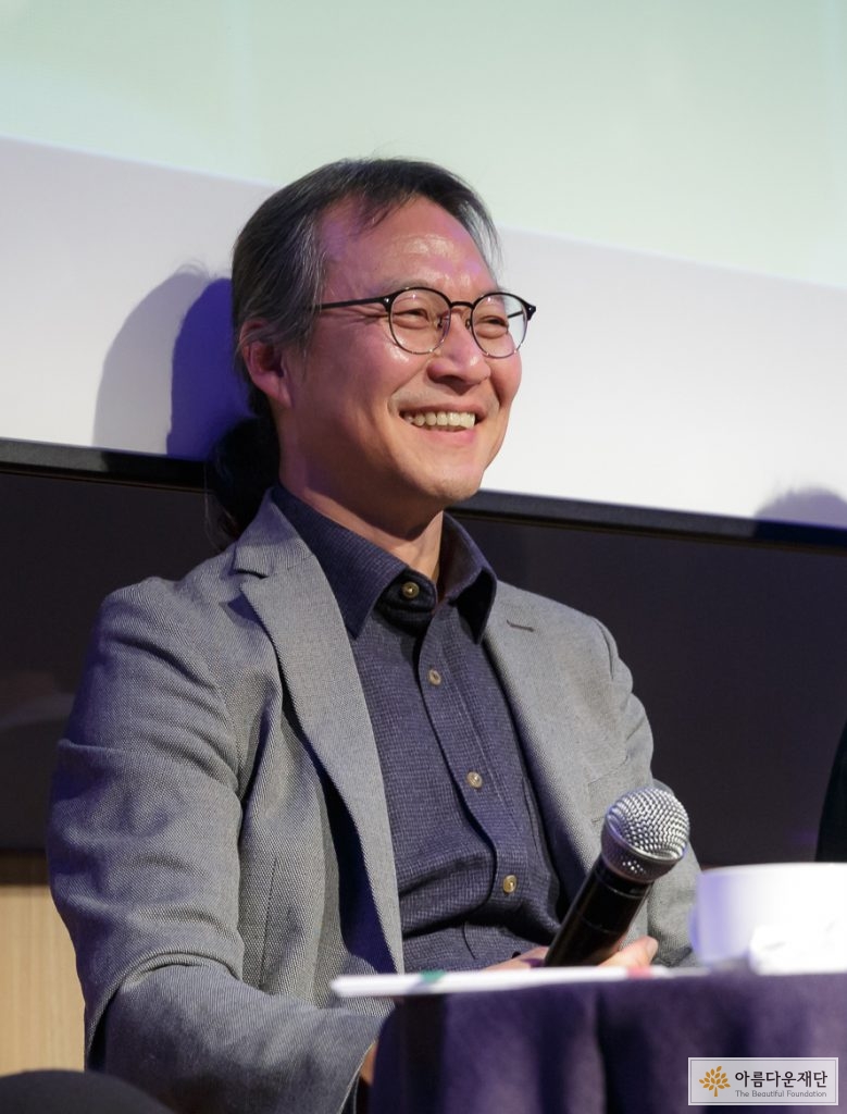 회색 자켓을 입고 안경을 쓴 남성이 손에는 마이크를 들고 앞을 바라보며 웃고 있음.