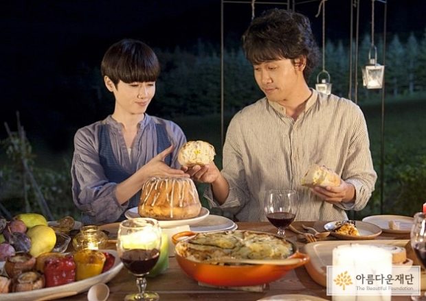 남자와 여자가 음식들로 차려진 식탁 앞에 앉아 있고 남자가 여자에게 빵을 건네주고 있다.